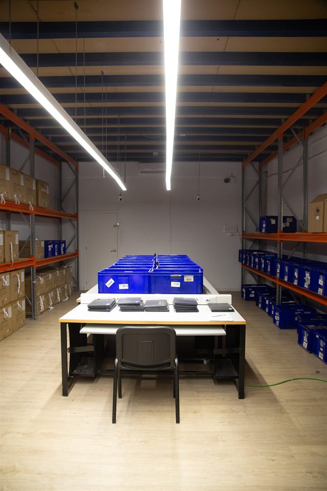 Sala con estanterías, cajas azules y una mesa con portátiles apilados para revisar.
