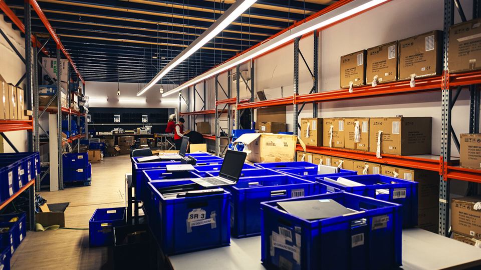 El almacén del taller: estanterías naranjas, cajas azules etiquetadas y portátiles en revisión.